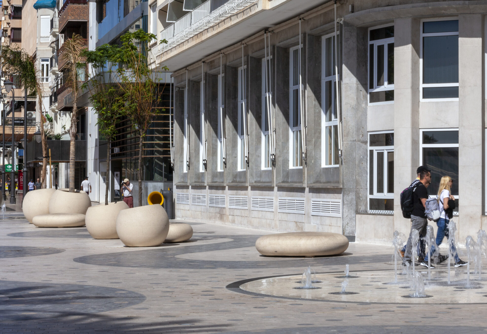 Mobiliario urbano de hormigón color beige, jardineras esféricas y bancos redondeados, fuente ornamental en el centro de la plaza